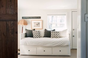 Un canapé blanc avec des draps à rayures, une lampe en osier, un tableau encadré et un panneau accroché au mur.