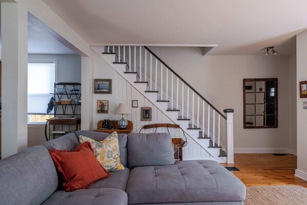 Un salon avec un canapé d’angle gris, un escalier et, au fond, un coin repas.