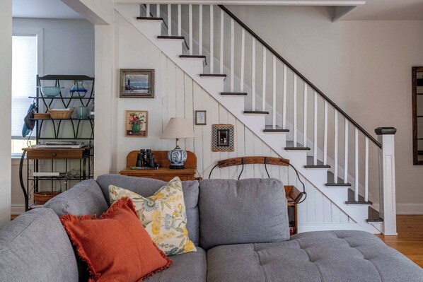 Un salon avec un canapé d’angle gris, une table basse en bois, un escalier avec des rampes blanches et un mur orné de tableaux et d’une lampe.
