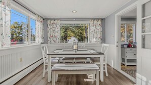 A dining area with a table and chairs, a window with floral curtains, and a view of trees outside. - Paws and Pines Retreat! (Ellsworth)