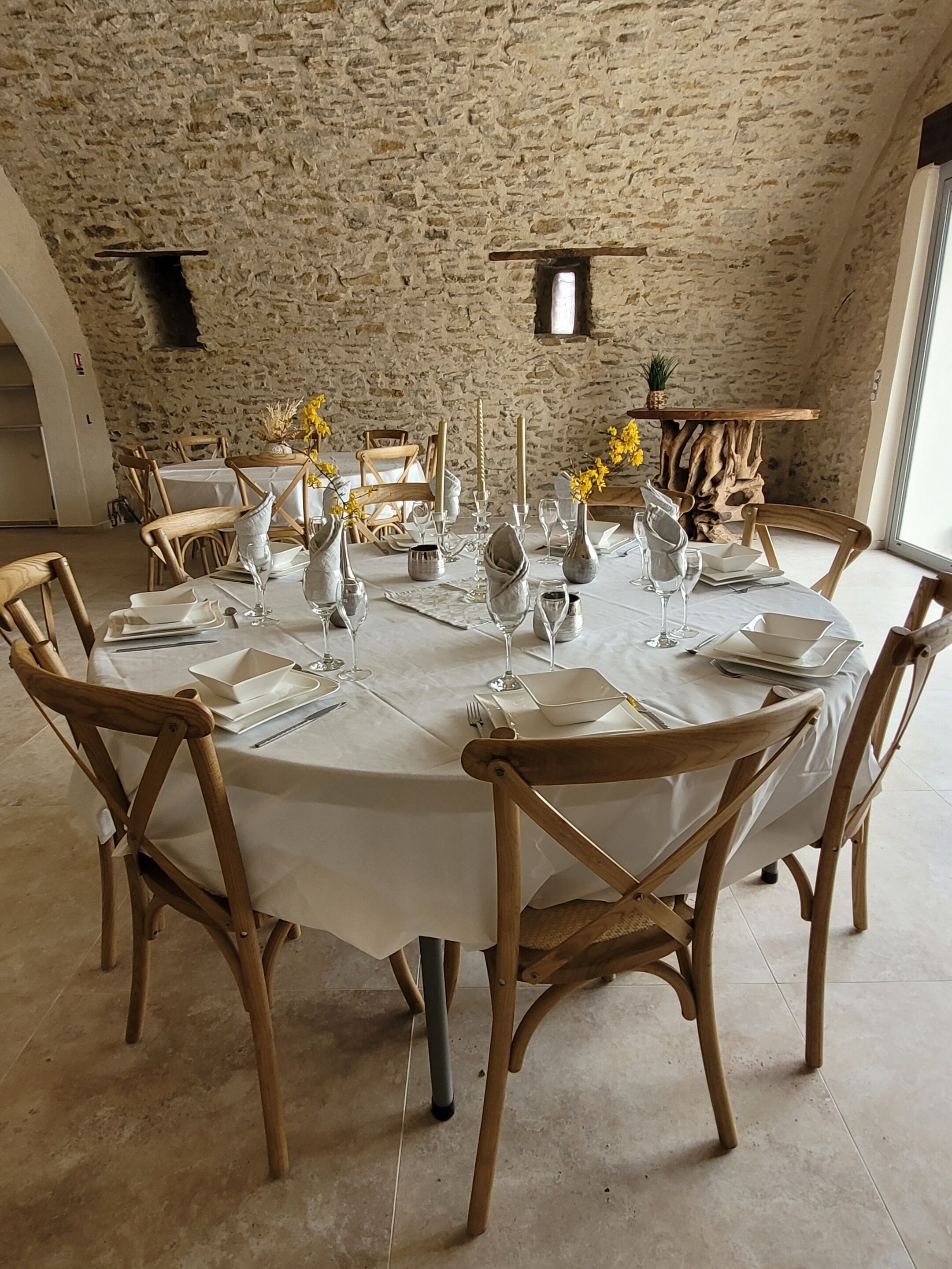 Mesa redonda de comedor con mantel blanco, rodeada de sillas de madera, dispuesta para una comida con velas y flores.