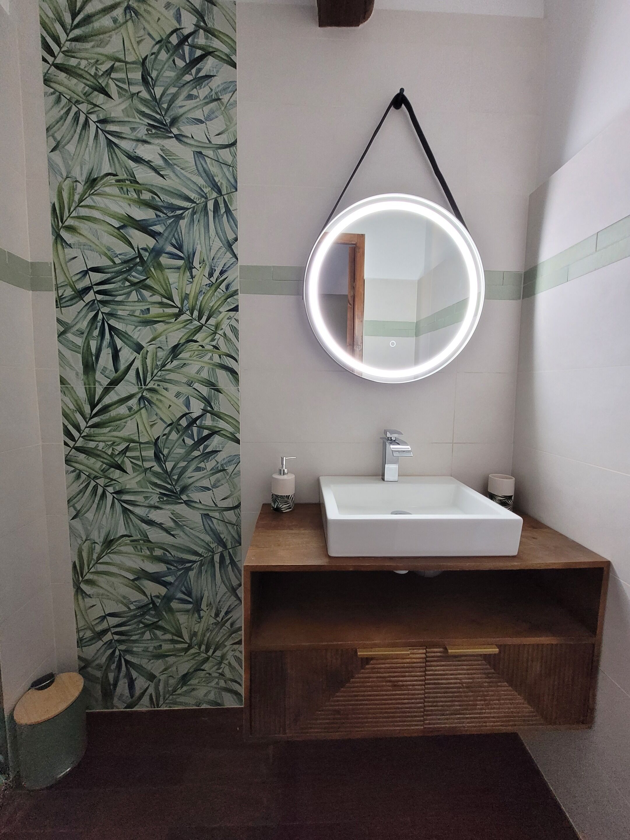 A bathroom with a wooden vanity, a white sink, a round mirror with a light, and a green leaf-patterned wall.