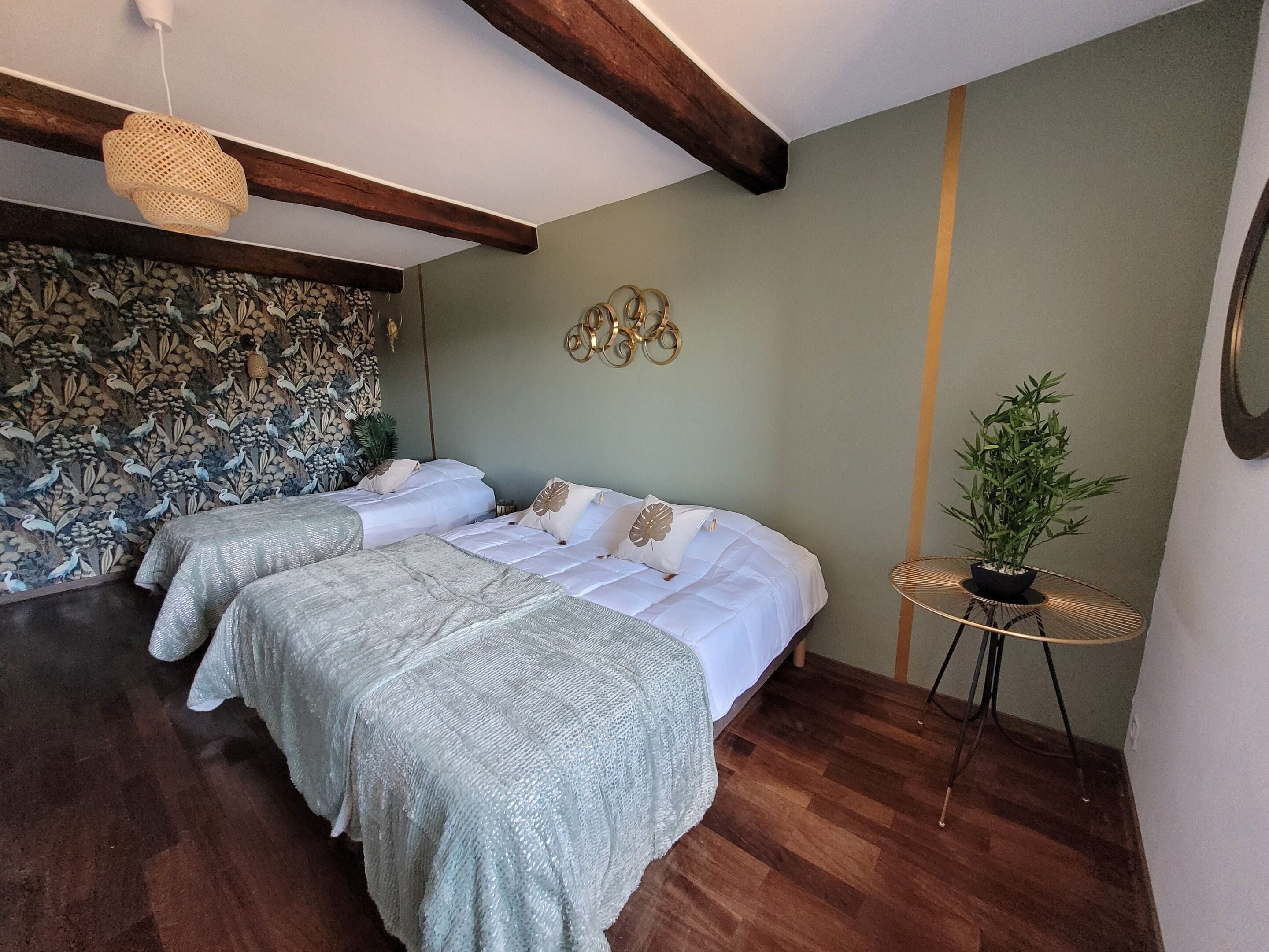 Una habitación con dos camas, una lámpara de fibra, un papel tapiz estampado, una pequeña mesa redonda con una planta y un adorno en la pared.