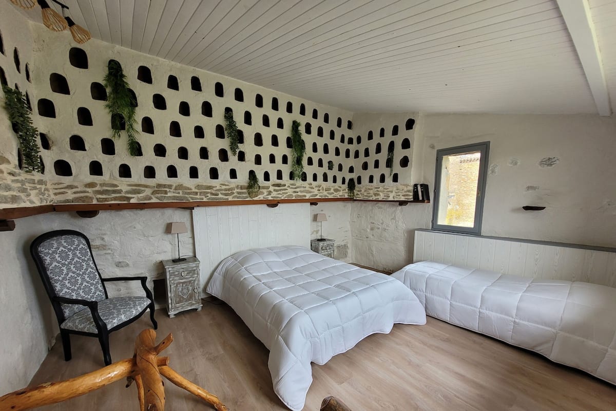 A bedroom with a stone wall, two beds, a chair, and a wooden bench.