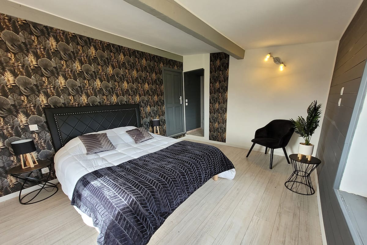 A modern bedroom with a large bed, a side table, a chair, and a potted plant.