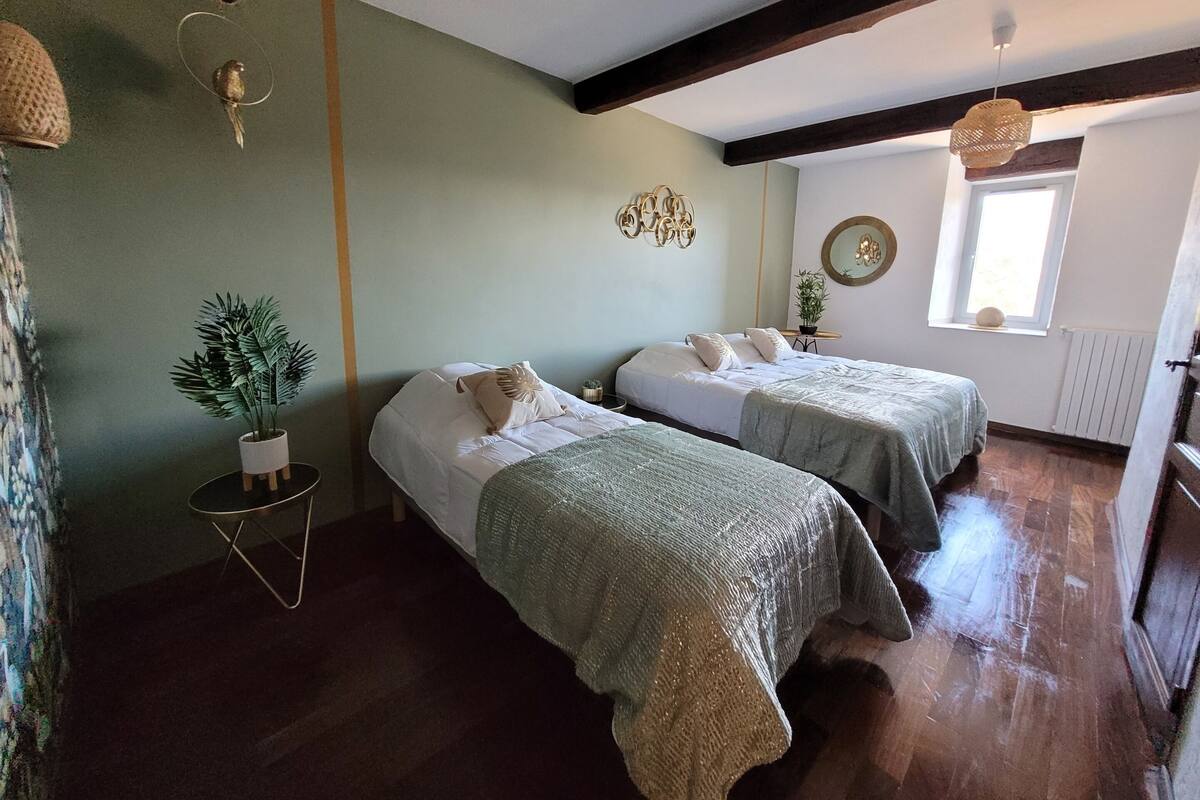 A room with two beds, a small table with a plant, a hanging light fixture, and a window with a view.