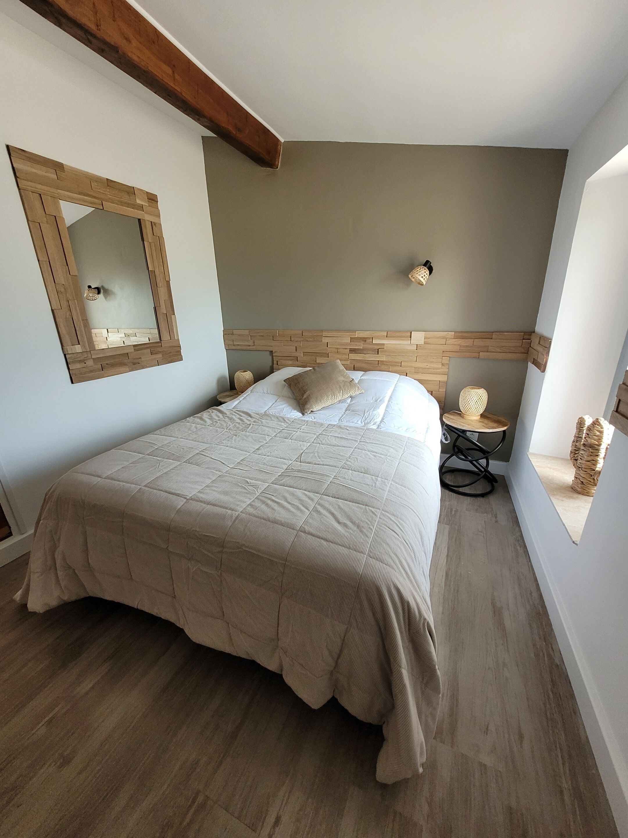 A bedroom with a large bed, wooden headboard, a wall-mounted mirror, and a small table with a lamp.