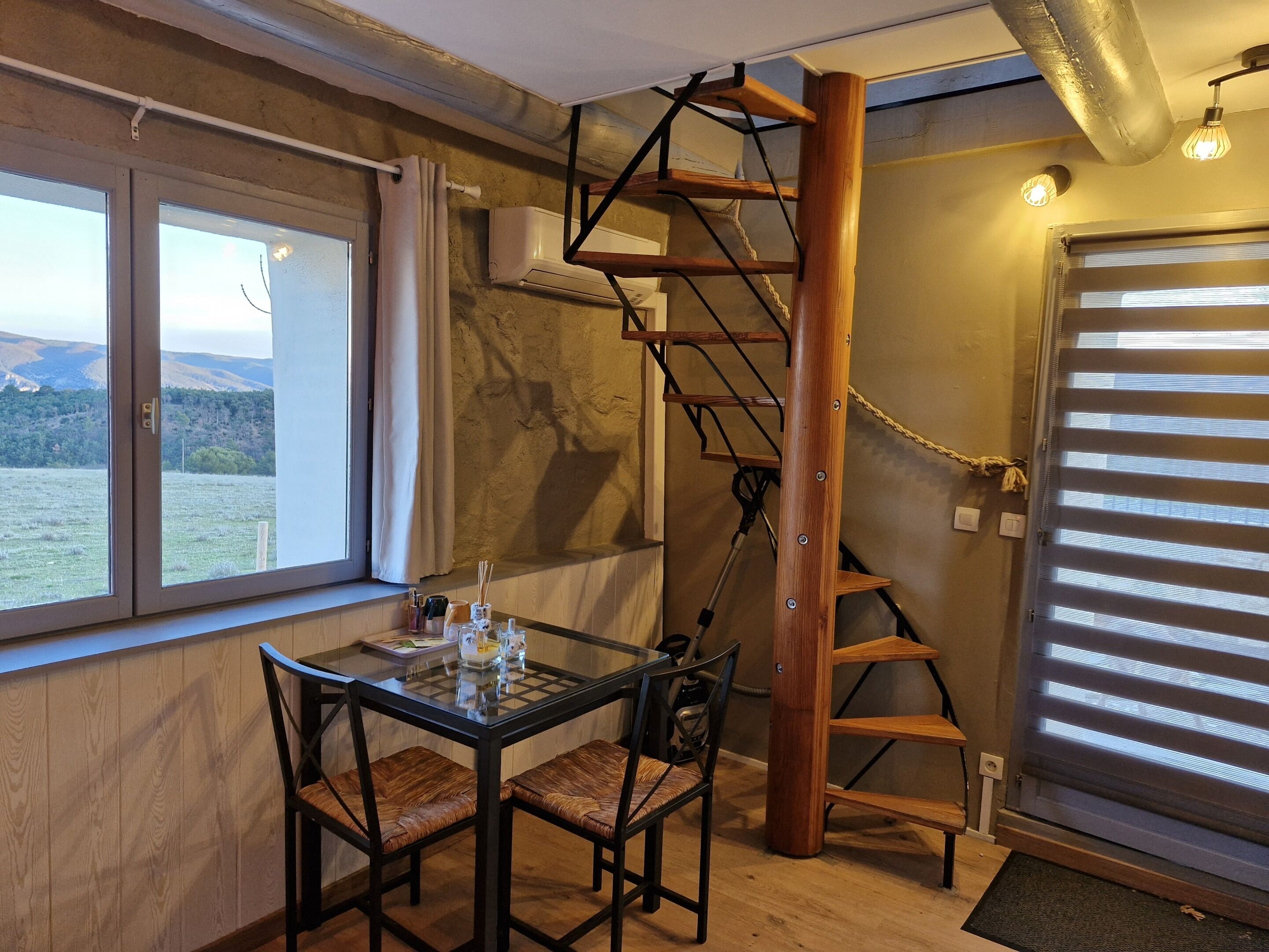 A dining area with a table and chairs, a wooden spiral staircase, a window with a view of the outdoors, and a door with blinds.