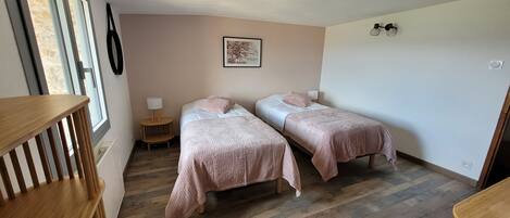 A bedroom with two single beds, wooden flooring, a small bedside table with a lamp, and a window with blinds.