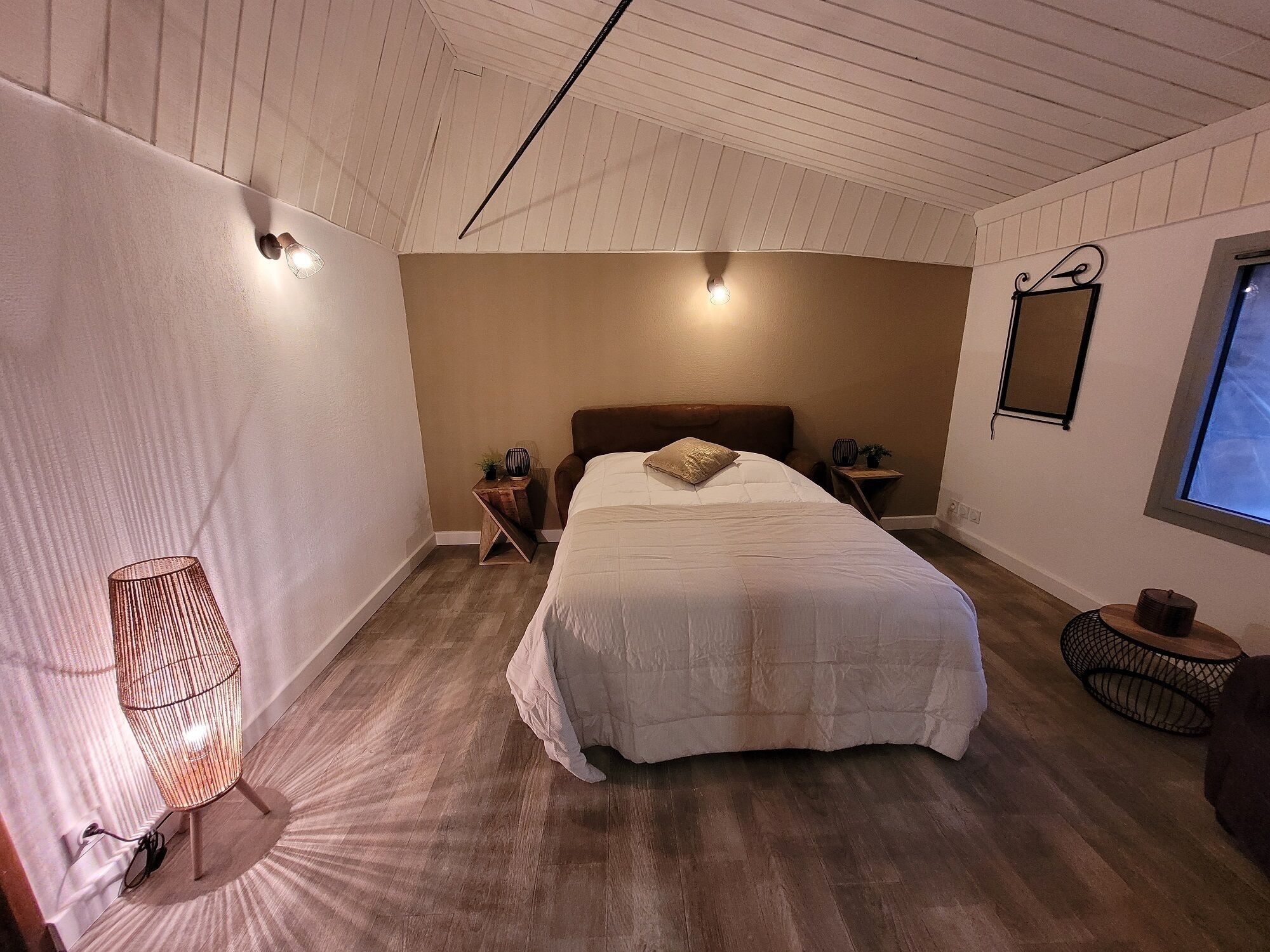 A bedroom with a large bed, a bedside table, a nightstand, a hanging light fixture, a small chair, and a window.
