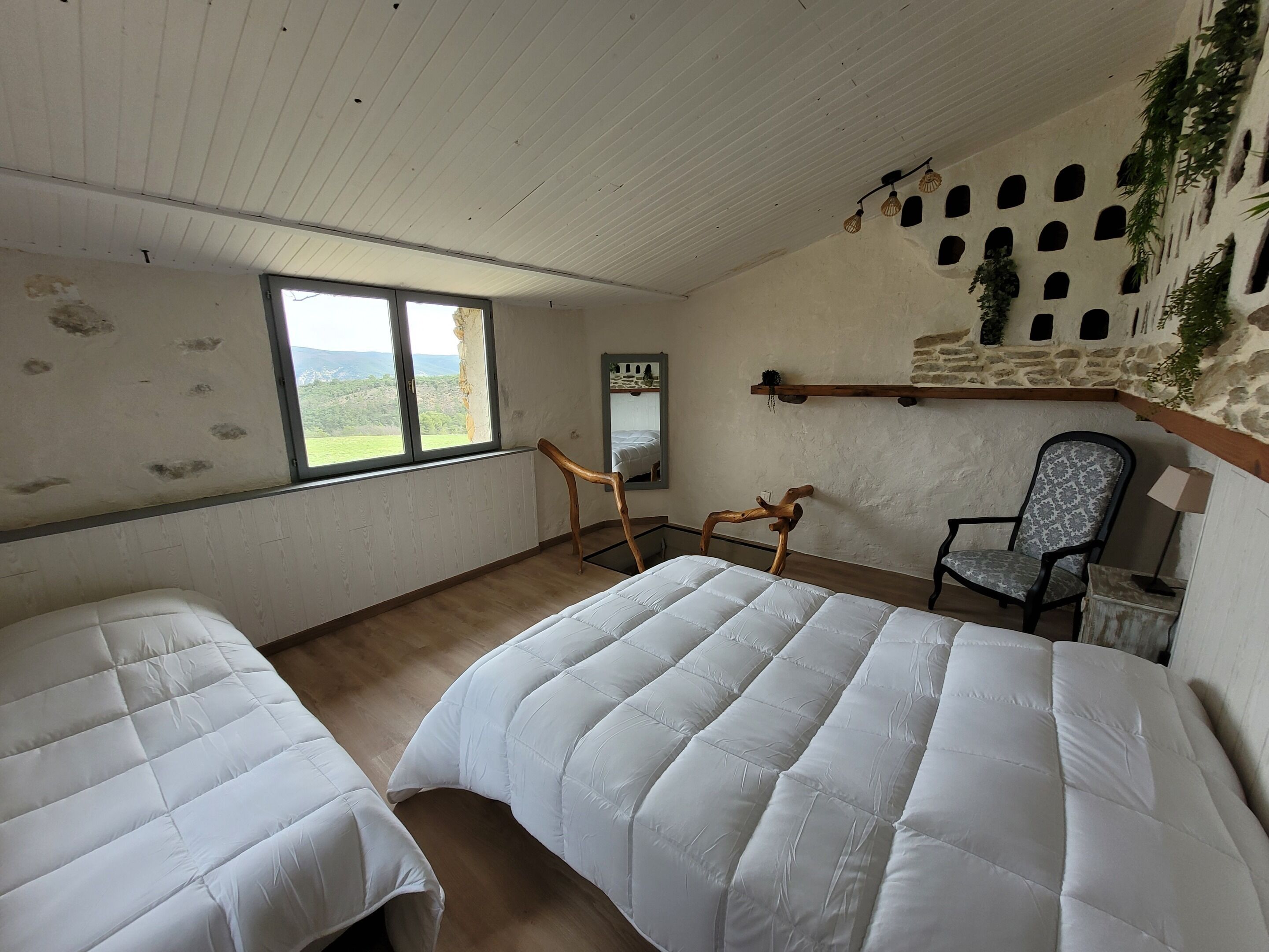 Habitación con dos camas, una silla de madera, un escritorio, un espejo y una ventana con vistas a la vegetación.