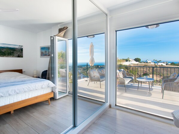 A bedroom with a large sliding glass door leading to a balcony with outdoor seating and a view of the sea.