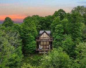 A cabin surrounded by dense forest, with a prominent balcony and a large window.