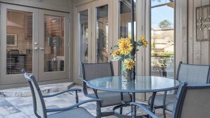Une terrasse avec une table ronde en verre et des chaises en métal, un vase rempli de fleurs jaunes et une porte-fenêtre coulissante.