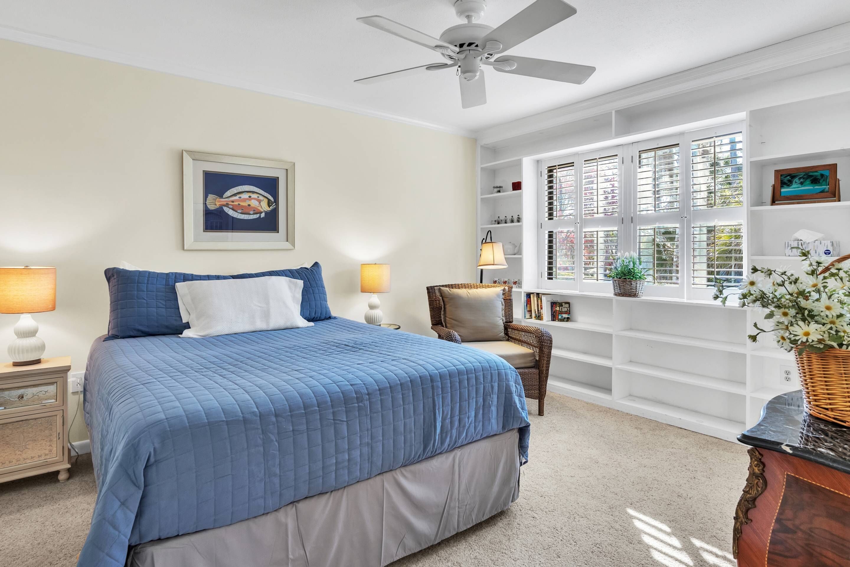 A bedroom with a large bed, a ceiling fan, a window with blinds, a nightstand, a chair, and a bookshelf.