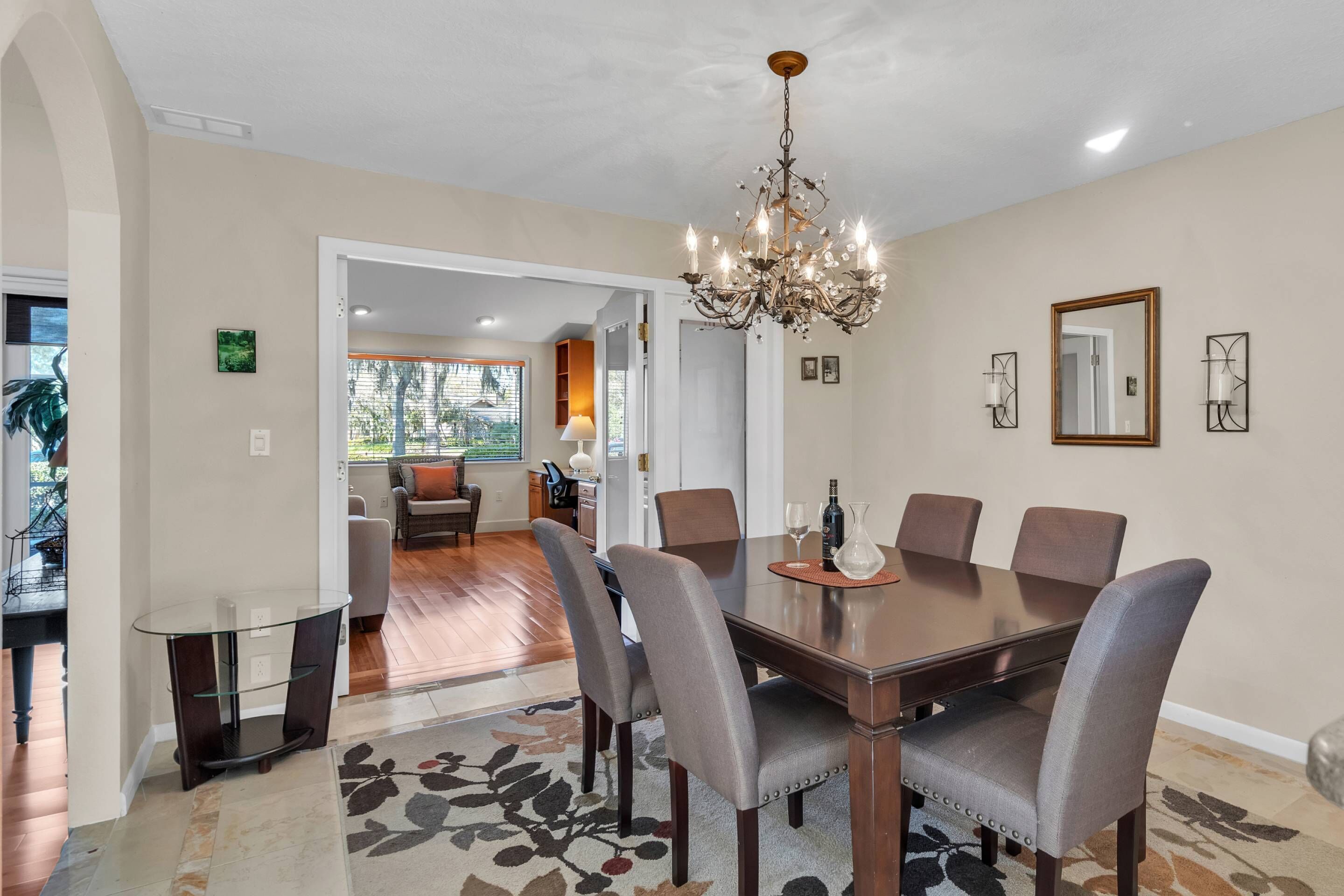 A dining area with a large wooden table, six upholstered chairs, a chandelier, and a patterned rug.