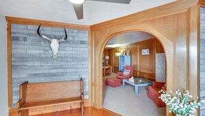 A room with a wooden bench, a ceiling fan, a stone wall with a bull skull, a red armchair, a blue table, and a vase with flowers. - Charming Farmington Home ~ 16 Mi to Aztec Ruins! (Farmington)