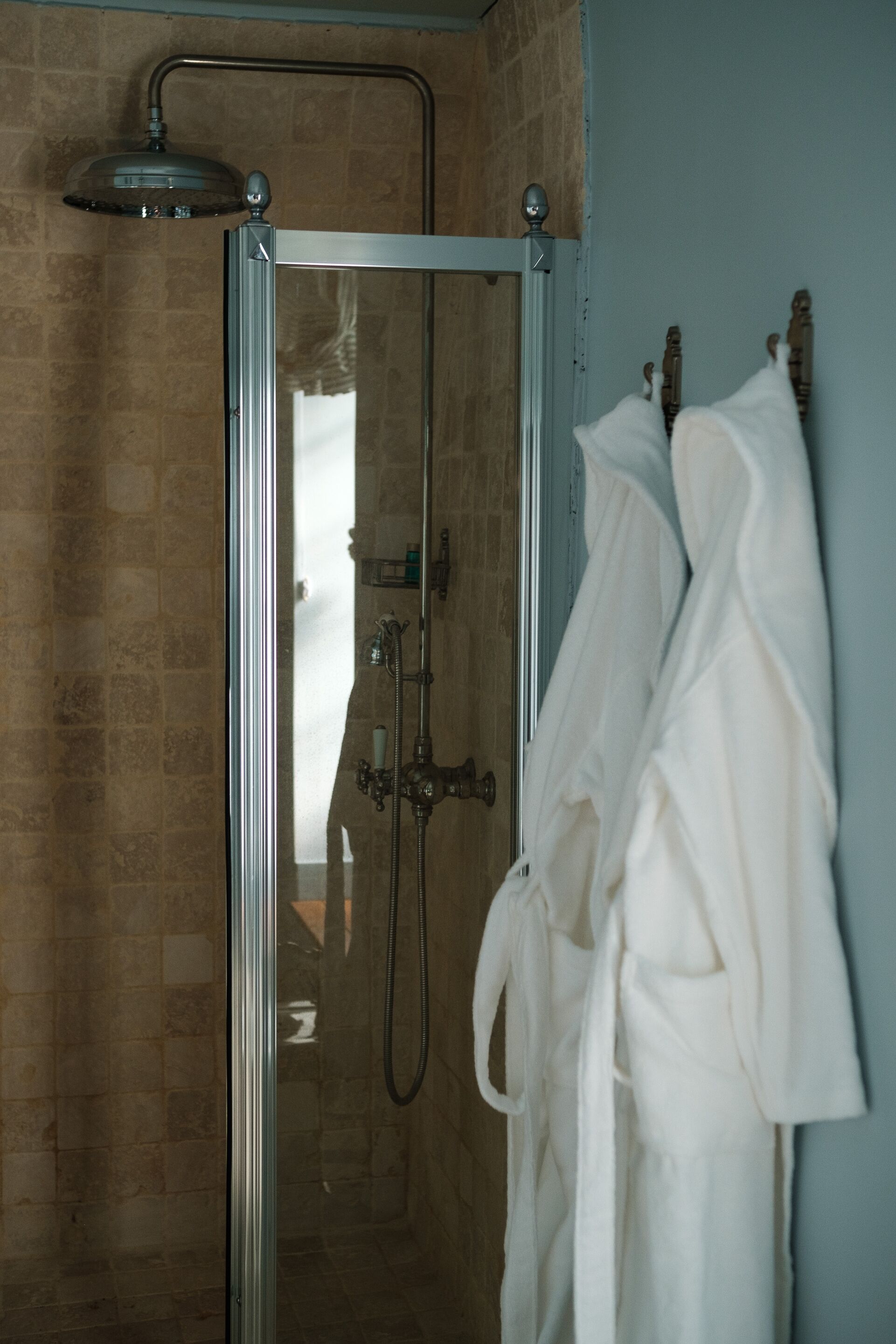 A glass-enclosed shower with a handheld showerhead, a towel rack with a white bathrobe, and a tiled wall.