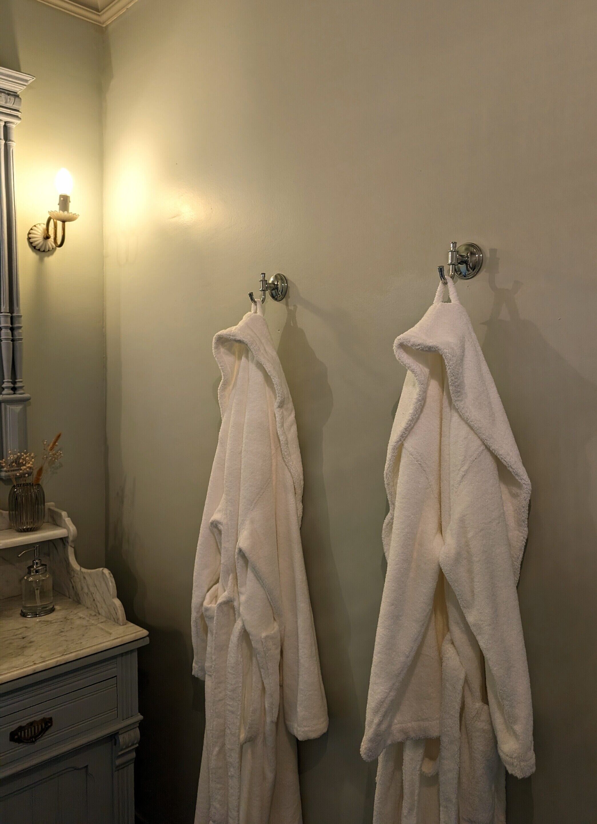 Dos batas blancas colgadas en ganchos en un baño con encimera de mármol y una lámpara en la pared.