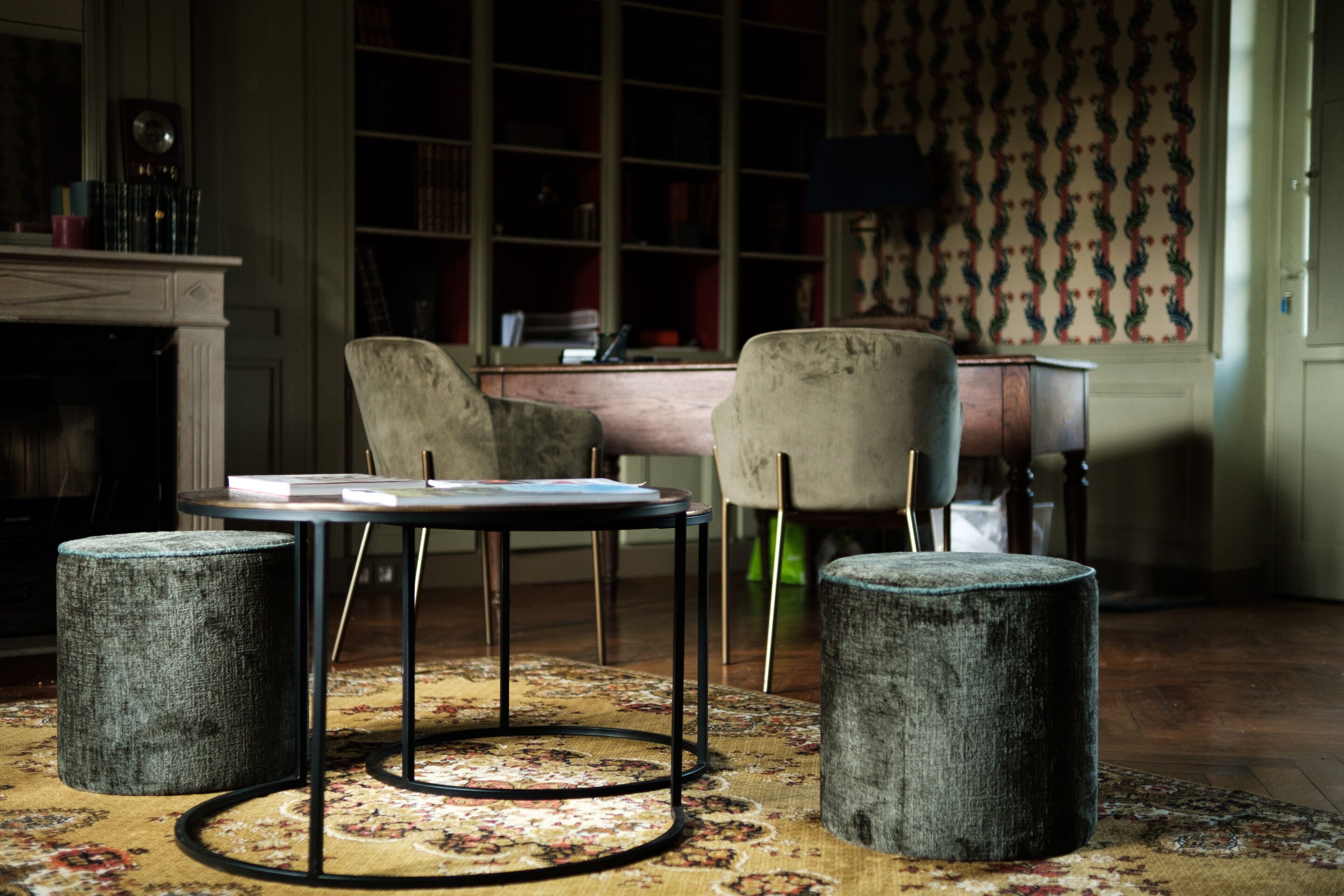 Une pièce comprenant une table ronde, deux poufs, une chaise, une bibliothèque et un tapis à motifs.