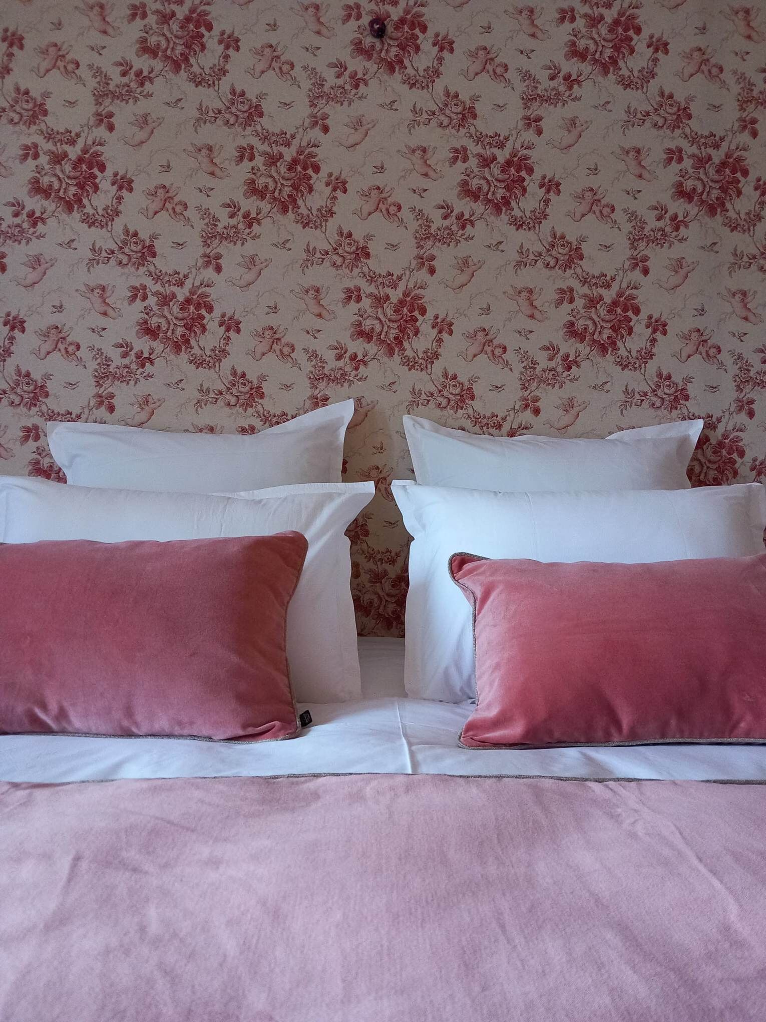 Uma cama com roupa de cama branca e rosa, papel de parede floral e uma parede branca lisa.