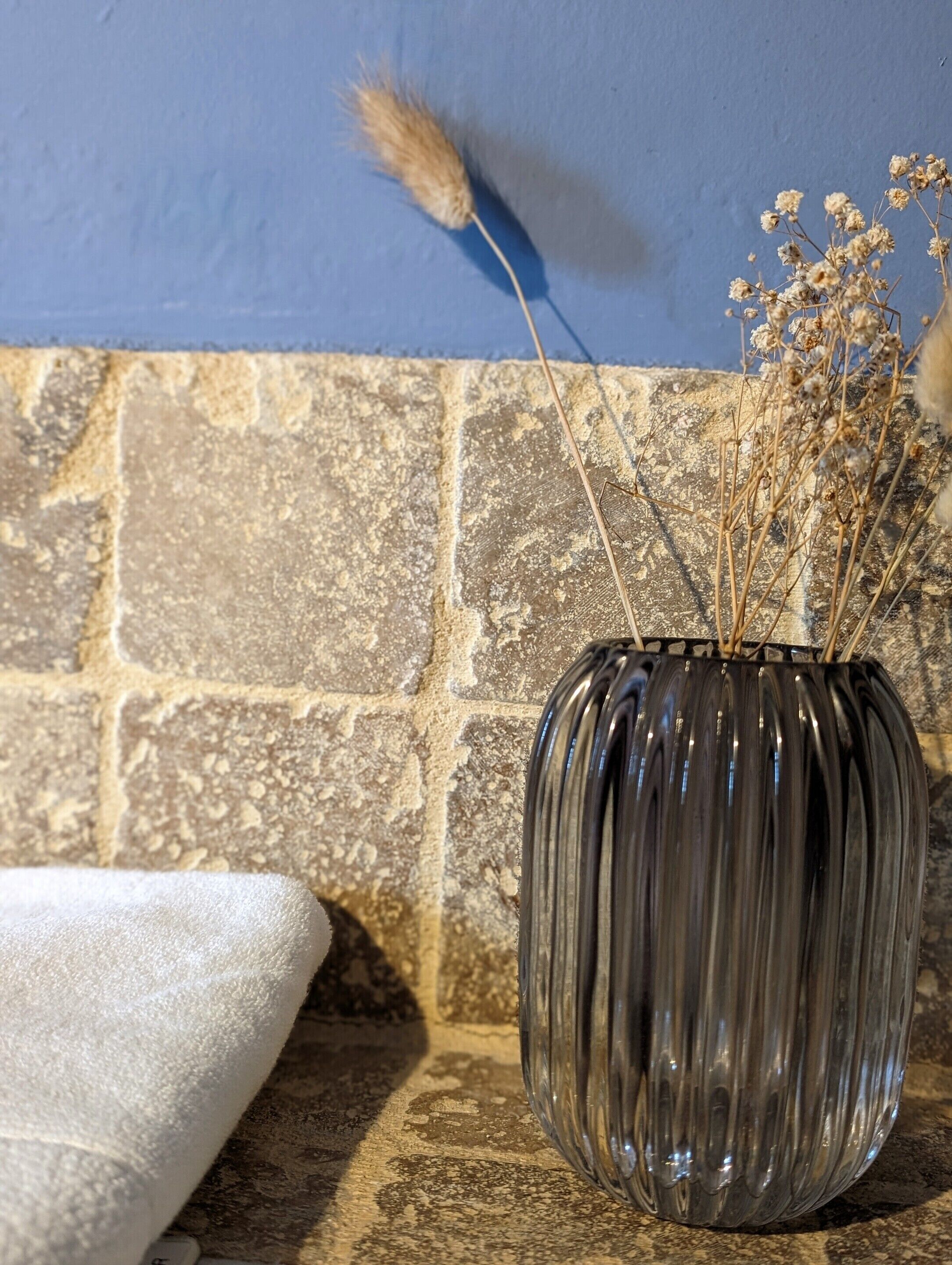 Un vase en verre transparent contenant des fleurs séchées, posé sur un mur en pierre à texture marquée.