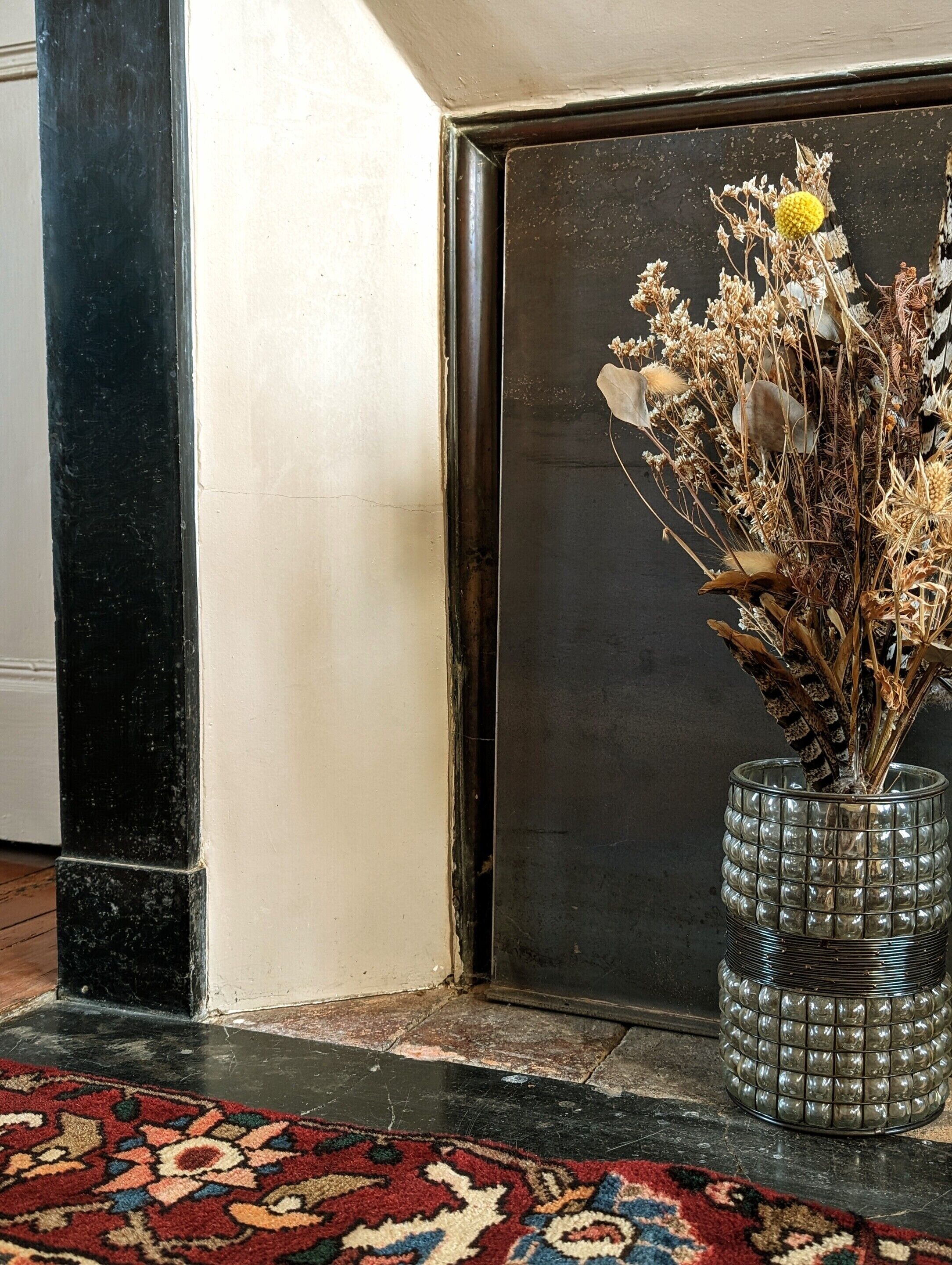 A corner of a room with a decorative vase on a patterned rug, a black door, and a white wall.