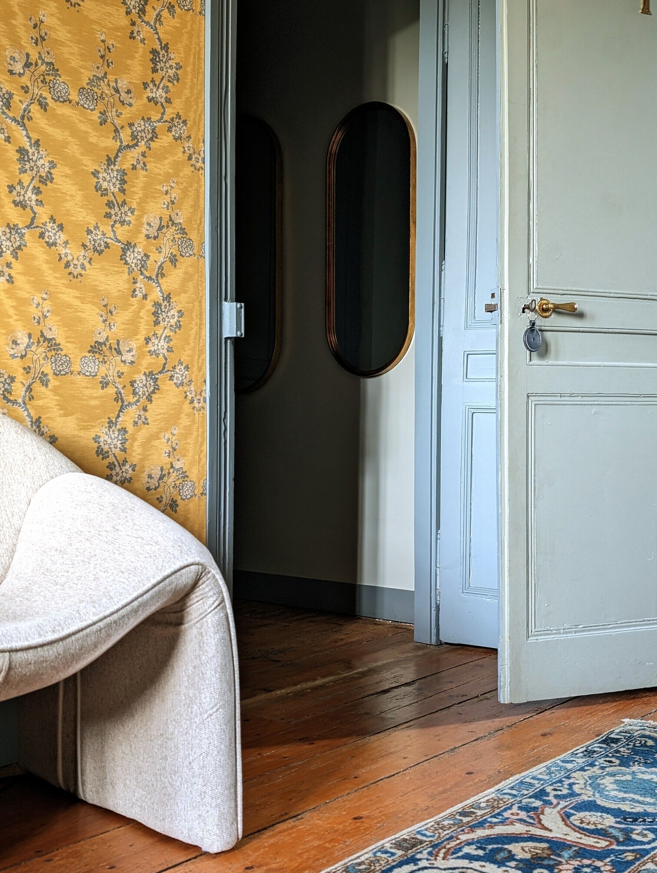 A room with a wooden floor, a white door with a brass handle, a patterned wall, and a small mirror.
