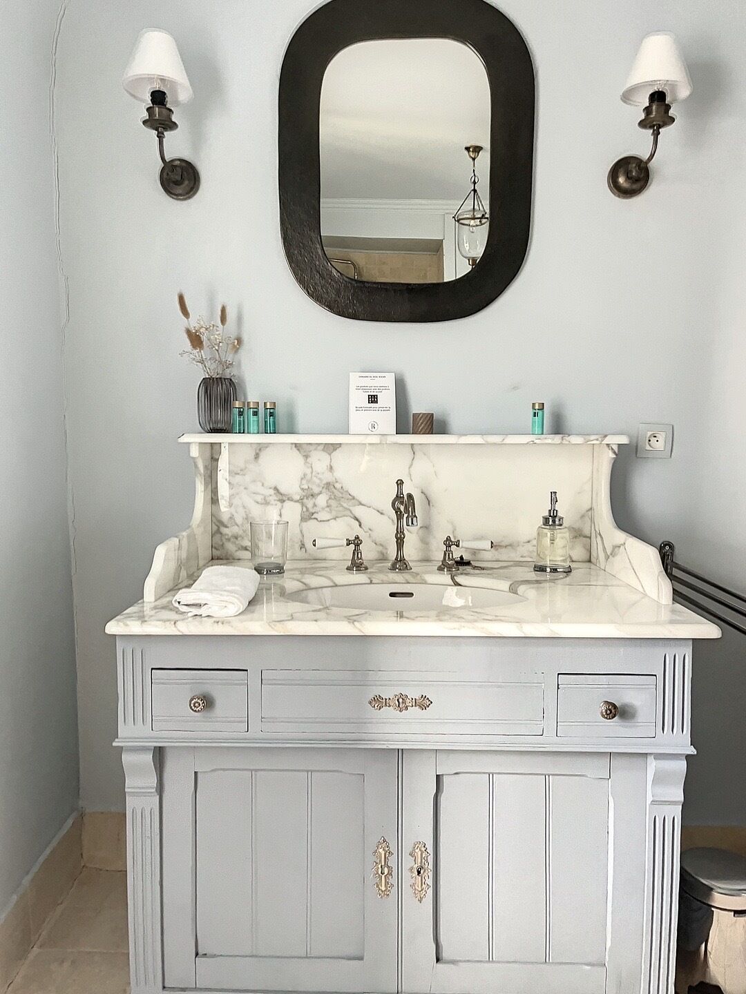 Une salle de bain avec un lavabo en marbre, deux appliques murales, un miroir et un petit vase contenant des fleurs séchées.