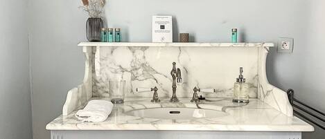 Une salle de bain avec un lavabo en marbre, deux appliques murales, un miroir et un petit vase contenant des fleurs séchées.