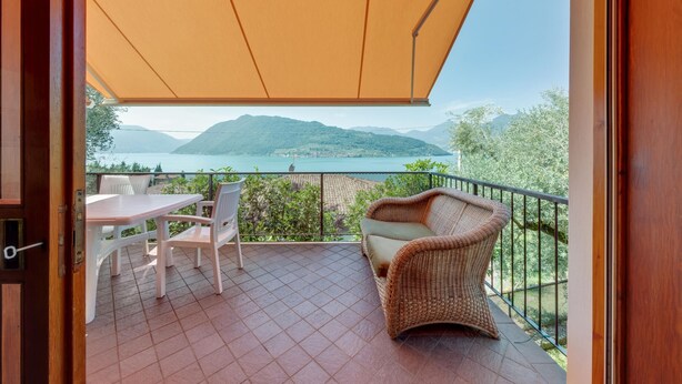 A balcony with a view of mountains and a lake, furnished with a table, chairs, and a wicker sofa. - Villa Olivella in Sale Marasino (Sale Marasino)