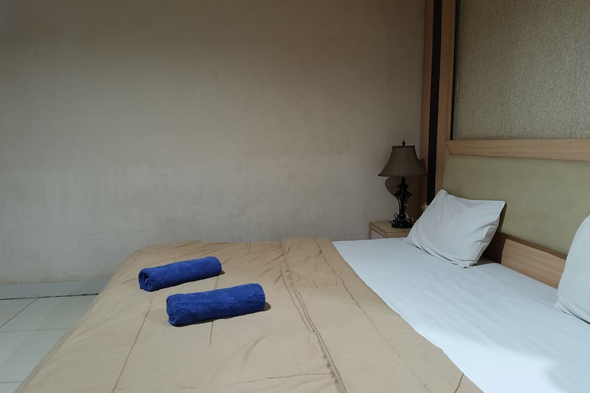 Habitación de hotel con cama, dos toallas enrolladas, mesita de noche con lámpara y cabecero con almohada.