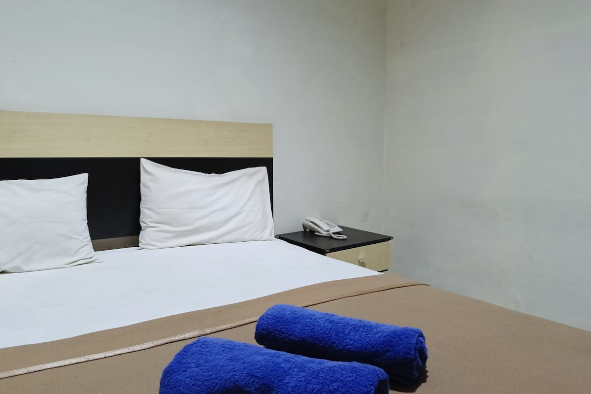 Una habitación de hotel con una cama, almohadas blancas, una mesita de noche negra con un teléfono blanco y dos toallas azules sobre la cama.