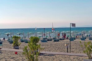 Una spiaggia con file di sedie a sdraio e ombrelloni verdi e bianchi, una barca a vela in acqua e un cartello in italiano.