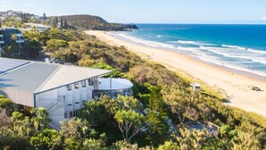 A coastal hotel with a beach view, surrounded by greenery and a hilly landscape.