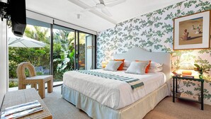 A bedroom with a large bed, a chair, a table with books, and a window with a view of greenery.