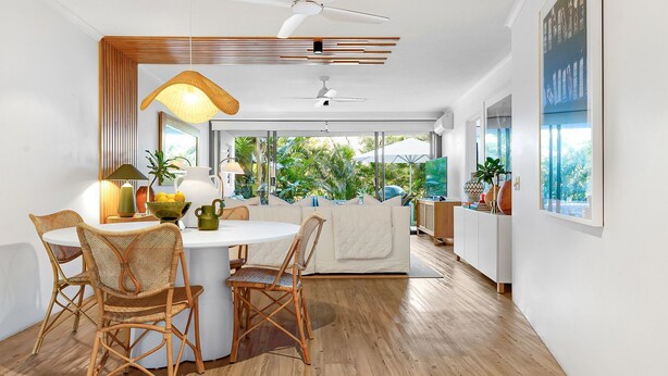 A modern kitchen with a dining area, featuring a round table, wicker chairs, and a view of the outdoors.