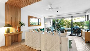A modern living room with a sofa, a wooden accent wall, a ceiling fan, and a view of an outdoor patio with furniture and greenery.