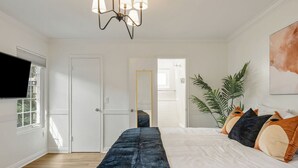 Un dormitorio con una cama grande, un televisor montado en la pared, una planta y una puerta que da a otra habitación.