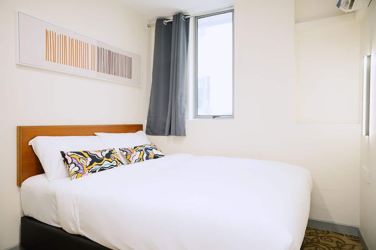 A neatly made bed with a wooden headboard, a colorful pillow, and a plain wall with a framed artwork above it.