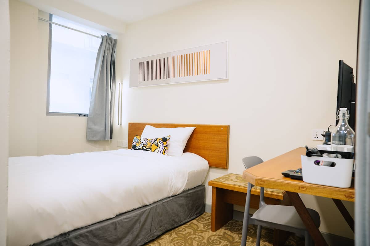 A hotel room with a bed, a desk, a chair, a window with curtains, and a wall-mounted artwork.