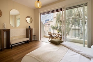 A bedroom with a bed, a bench, a round mirror, and a balcony with a table and chairs. - MAMAD - Seabreeze Serenity Haven by Sea N' Rent (Tel Aviv)