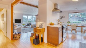 A modern kitchen with wooden cabinetry, a central island, and a dining area with a view of the outdoors. - Four Bears by AvantStay | Walk to Beach, Hot Tub, Ocean View (Arch Cape)