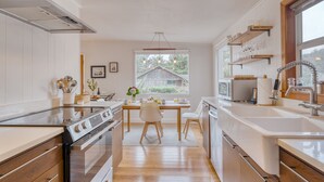 A modern kitchen with wooden cabinets, a central island, and a view of a house through a window. - Four Bears by AvantStay | Walk to Beach, Hot Tub, Ocean View (Arch Cape)