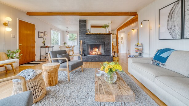 A spacious living room with a fireplace, a sofa, a coffee table with flowers, and a dining area in the background. - Four Bears by AvantStay | Walk to Beach, Hot Tub, Ocean View (Arch Cape)