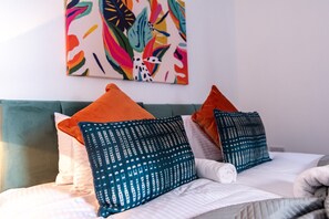 Ein Bett mit weißer Bettwäsche, blaue Kissen in Türkis- und Orangetönen und ein farbenfrohes abstraktes Gemälde an der Wand.
