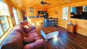 A cozy cabin interior with a wooden floor, a sofa, a dining table, a kitchen area, and a TV mounted on the wall.