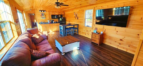 Interior acogedor de una cabaña con piso de madera, un sofá, una mesa de comedor, una zona de cocina y un televisor montado en la pared.