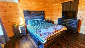 A wooden cabin bedroom with a bed, dresser, nightstand, and a television.