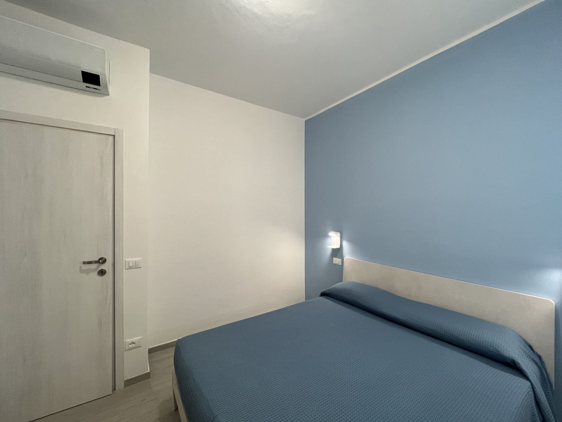 Un dormitorio con una cama individual, una puerta, un aire acondicionado de pared y un interruptor de luz.